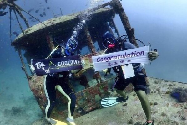 Two scuba divers underwater near a shipwreck, holding a sign that says 'Congratulations.'