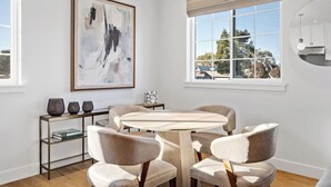 A modern dining area with a round table and four chairs, a large abstract painting on the wall, and a window with a view of trees and houses. - 2 bedroom - WiFi, AC, garaged off-street parking with EV Charger. (Solvang)
