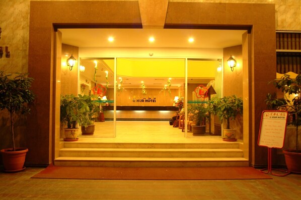 The entrance of a hotel with a reception desk, potted plants, and a signboard.