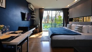 A modern hotel room with a large bed, a desk with a chair, a TV, and a view of greenery outside.