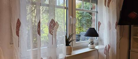 Een kamer met een raam voorzien van dunne gordijnen met een bladmotief, een witte lamp op de vensterbank en een televisie aan de muur.