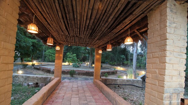 A covered walkway with brick walls and hanging lights, leading through a garden area with trees and a central water feature. - Mridu Kishore Resort (Bichhiya)
