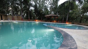 A large, curved swimming pool with clear water, surrounded by a stone deck and trees. - Mridu Kishore Resort (Bichhiya)