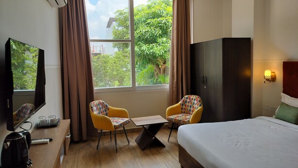 A hotel room with a bed, two chairs, a small table, a TV, a wardrobe, and a window with curtains. - Saffron Inn  (Greater Noida)