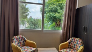 A room with two yellow chairs with colorful cushions, a small table, and a window with a view of trees and buildings. - Saffron Inn  (Greater Noida)