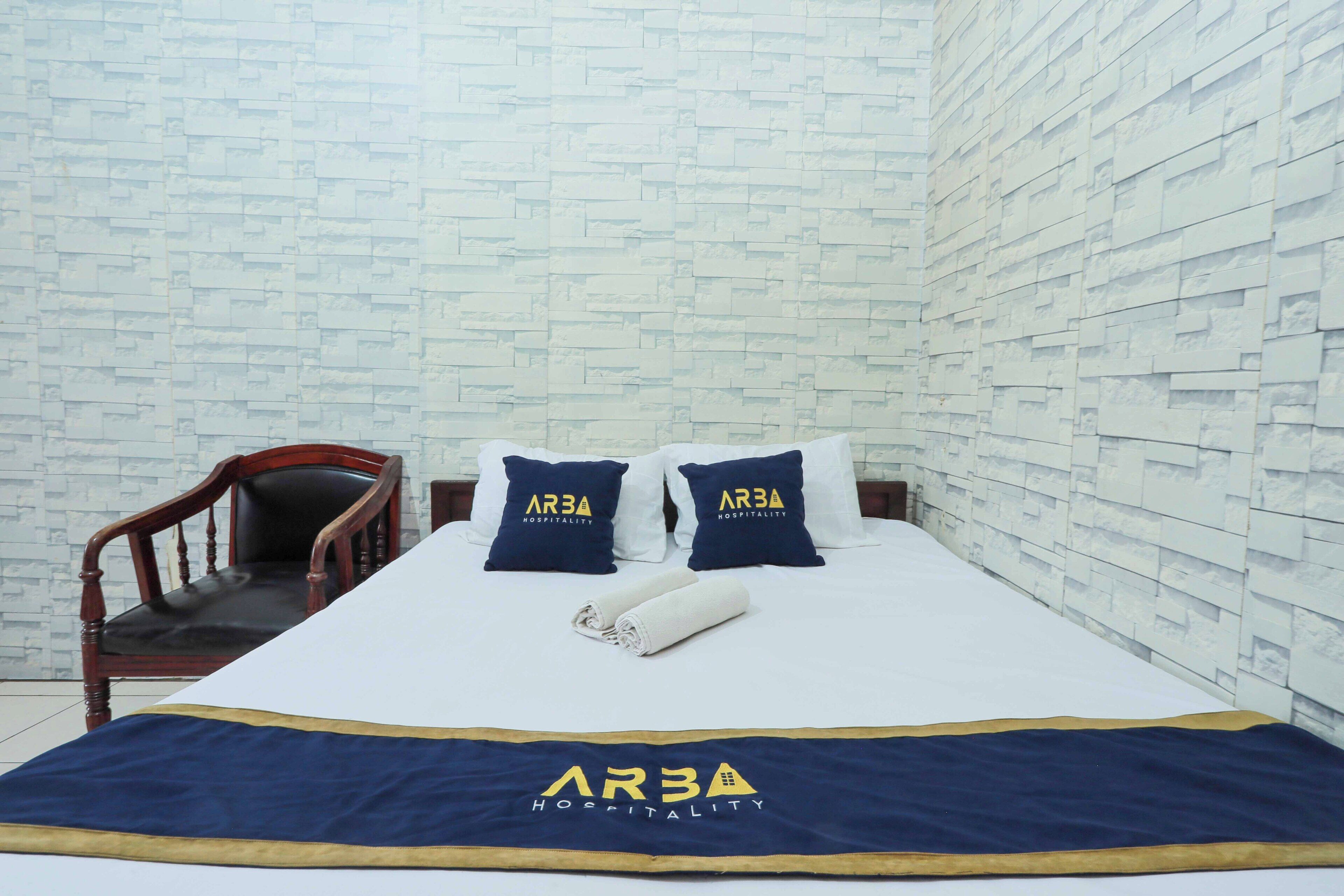 A hotel room with a neatly made bed, two pillows with the hotel's logo, a wooden chair, and a white brick wall.