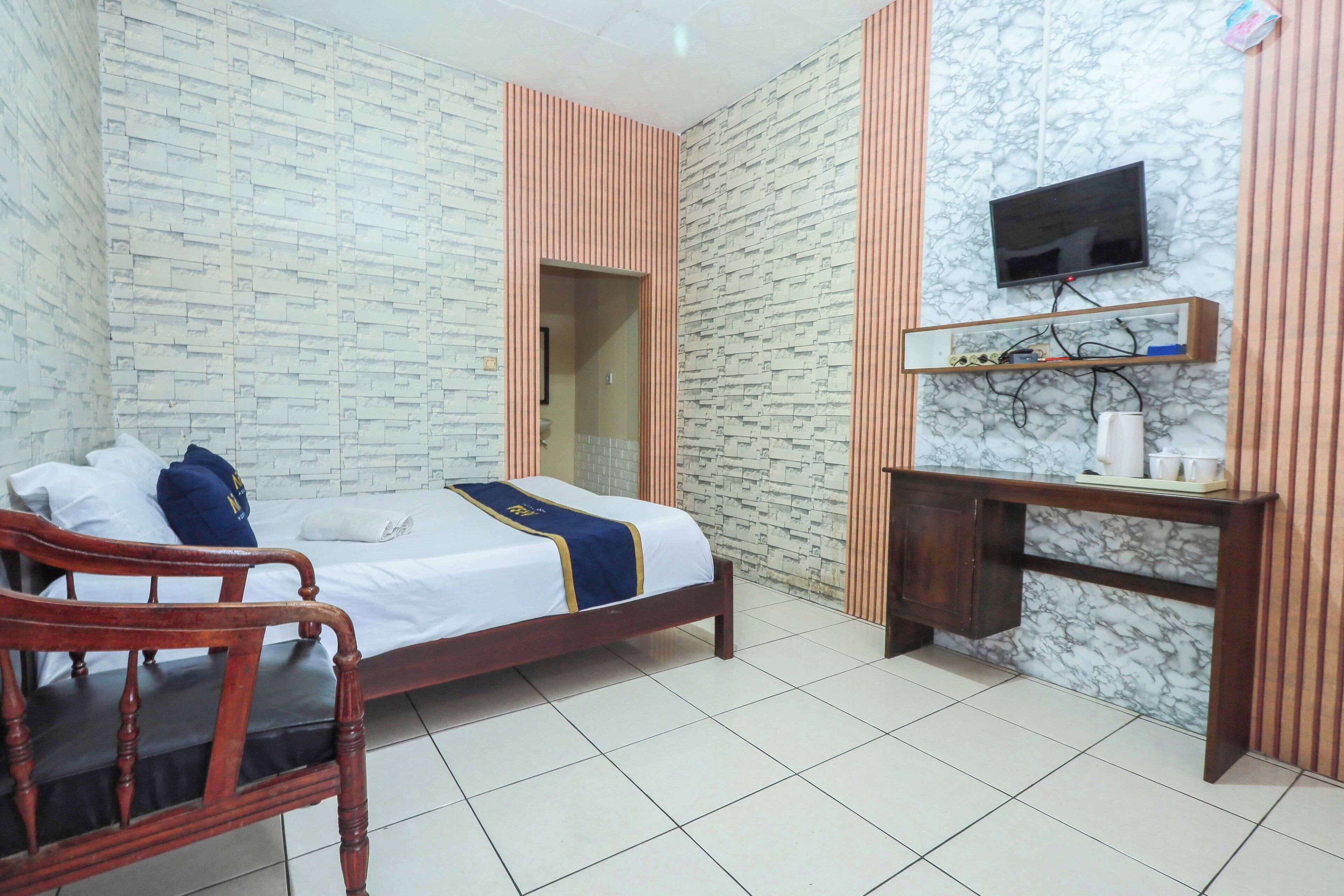 A hotel room with a bed, a wooden chair, a desk with a TV, and a marble-patterned wall.