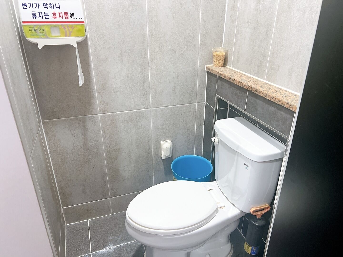 Un baño pequeño con un inodoro blanco, un cubo azul y un porta papel higiénico en la pared.