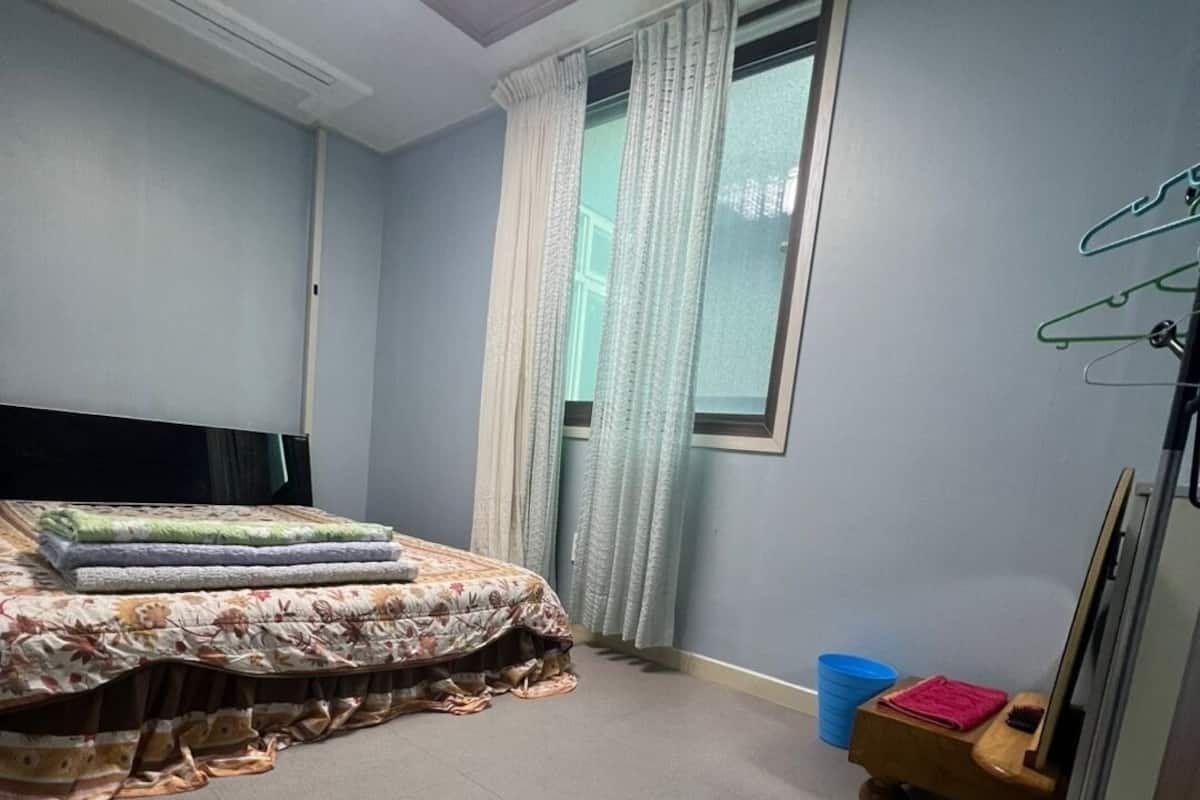 A bedroom with a bed, a window with curtains, a small table with items, and a ceiling light.