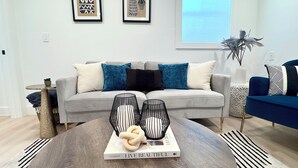 A modern living room with a grey sofa, blue and white cushions, a round coffee table with a book titled 'LIVE BEAUTIFUL', and a decorative plant. - Charming Madrona Stay • Walk to Lake & Cafes, Shops, A/C • Parking (Seattle)