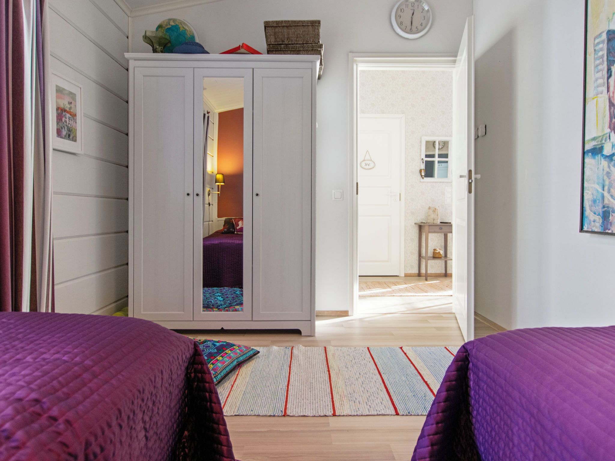 Une chambre avec deux lits, une armoire, une horloge et une porte donnant sur une autre pièce.