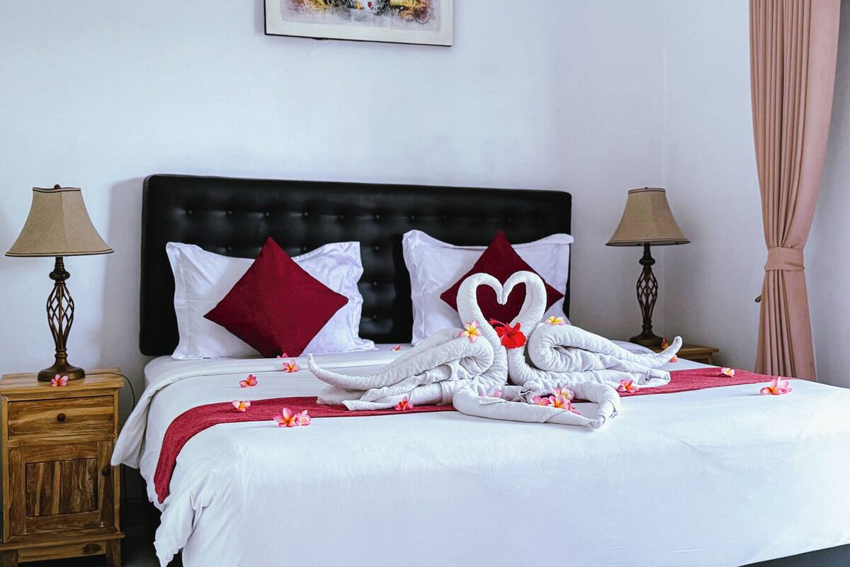A neatly made bed with white and red pillows, a heart-shaped towel arrangement, and a floral decoration on the nightstand.
