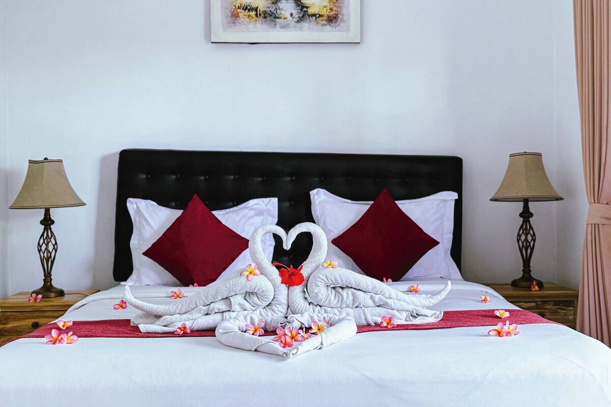 A neatly made bed with a towel swan centerpiece, red pillows, and a red throw blanket.