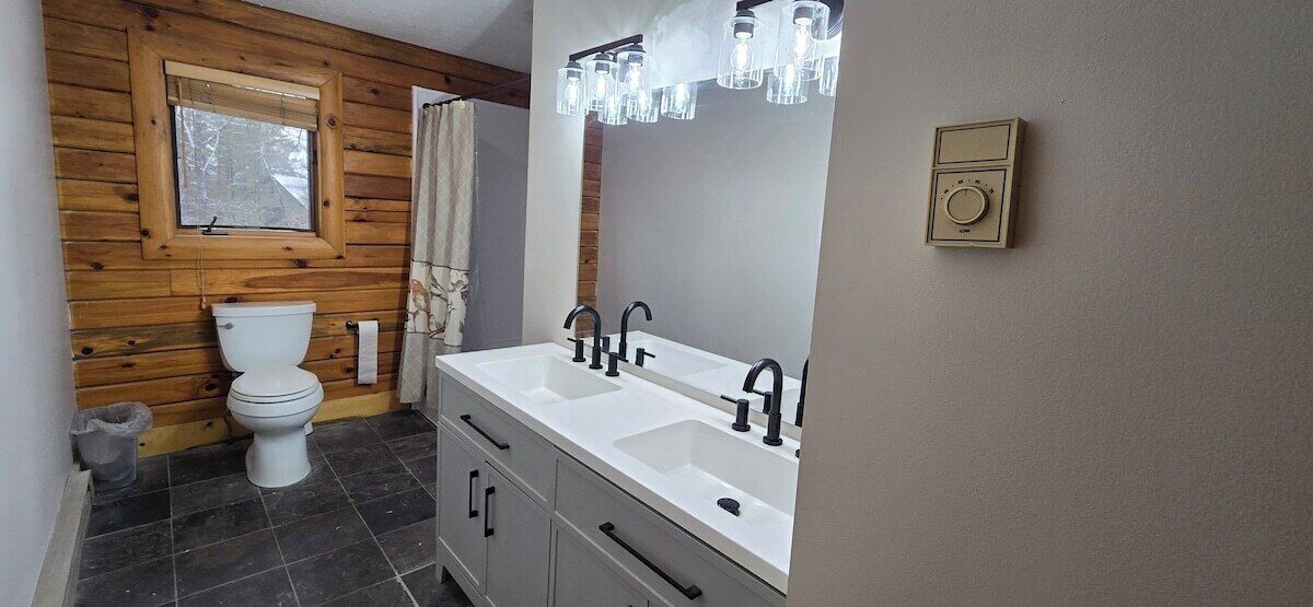 A bathroom with a wooden wall, a window, a white toilet, a double sink with black faucets, and a mirror with light fixtures above.