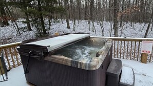 Un jacuzzi en una terraza de madera con barandilla metálica, rodeado por un bosque nevado.