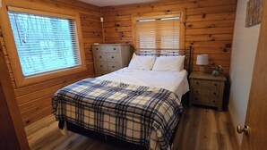 Un dormitorio con pared de paneles de madera, cama con colcha escocesa, mesita de noche con lámpara y cómoda.
