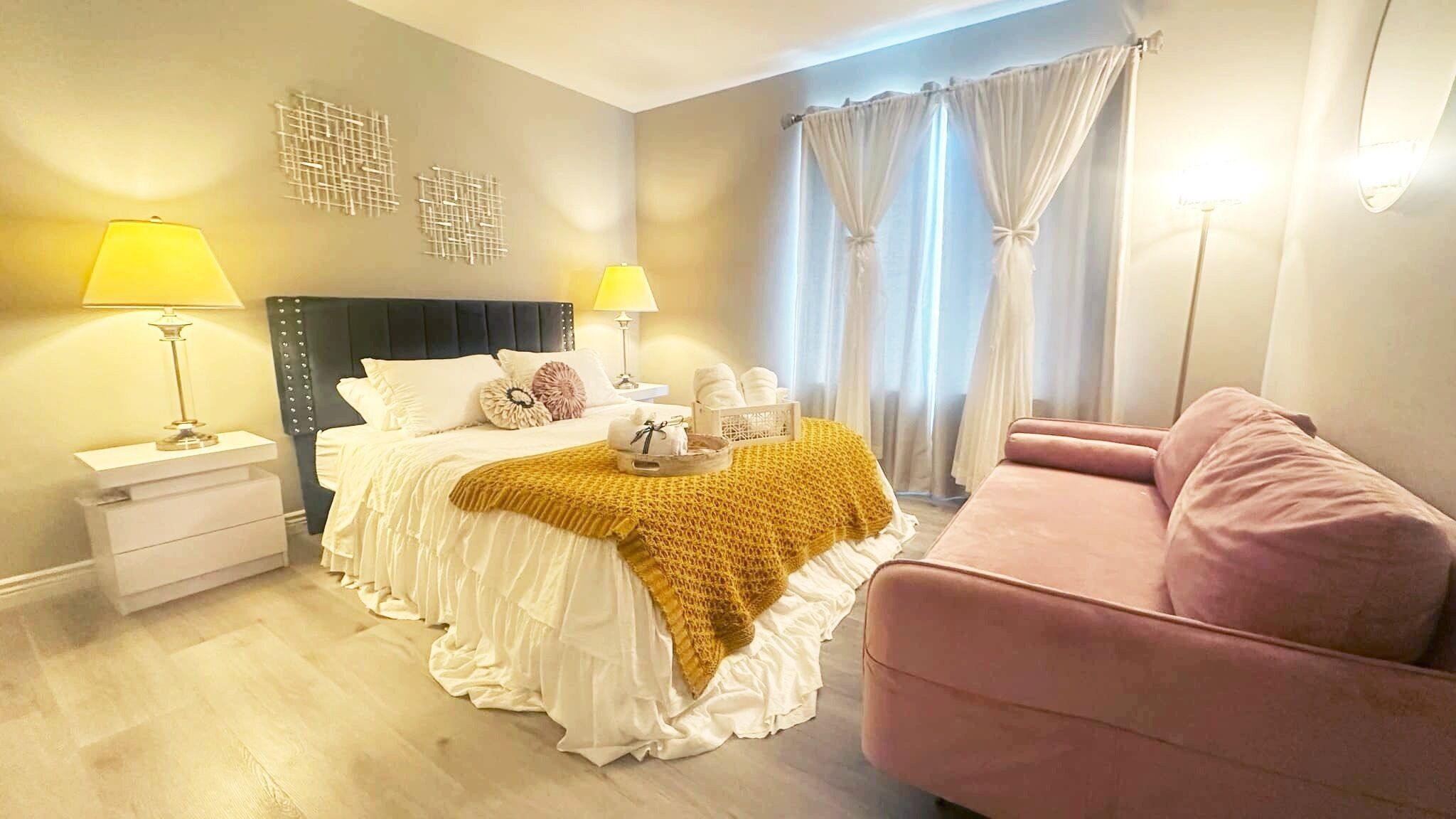 A bedroom with a bed, bedside tables, a pink chair, and a window with curtains.