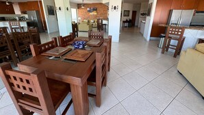Un'ampia zona giorno con tavolo da pranzo per sei persone, circondato da sedie in legno. Si intravede sullo sfondo una cucina con elettrodomestici moderni e mobili in legno.