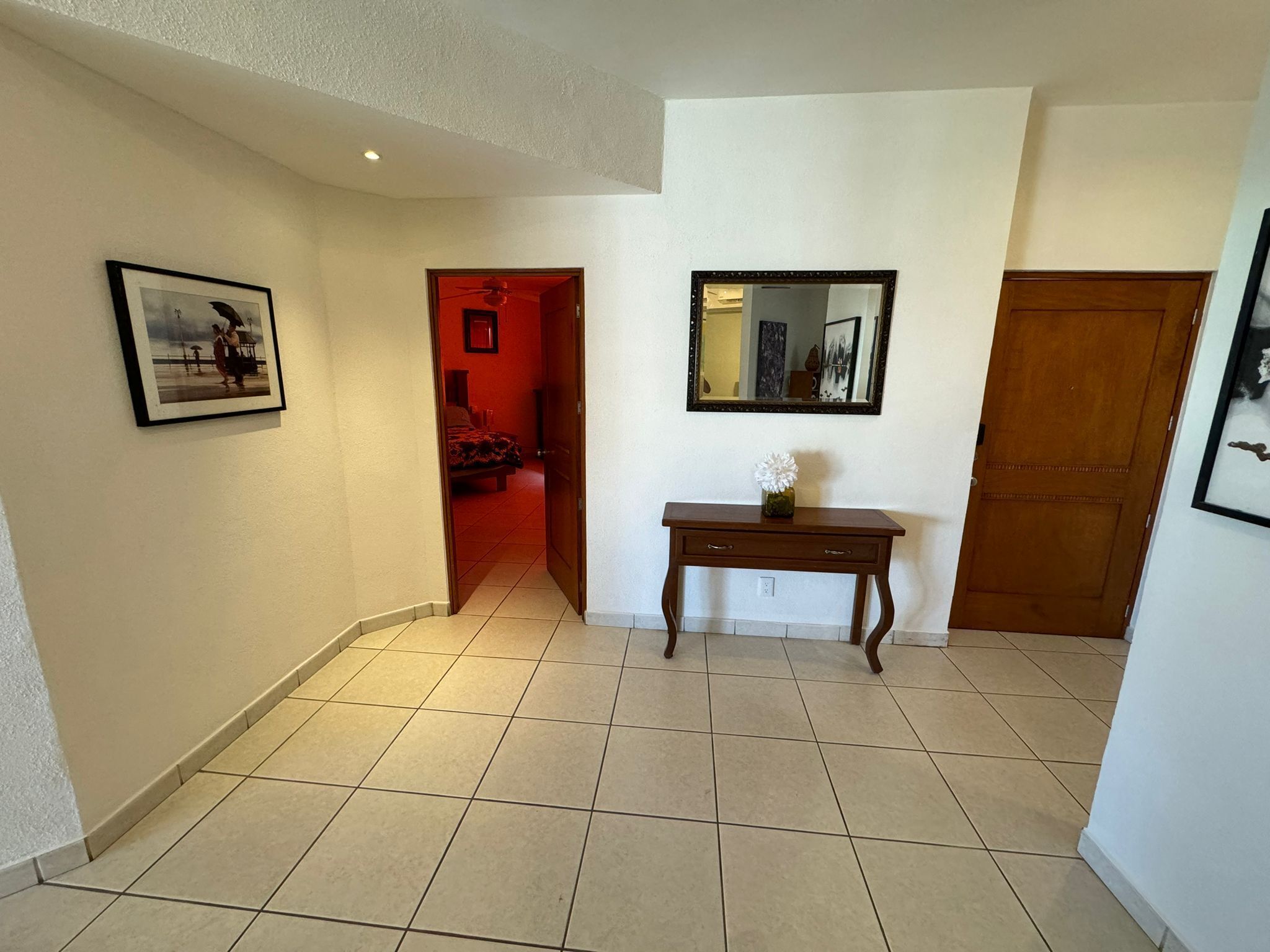 A hallway with a tiled floor, a wooden door, a mirror, and a small table with a vase.