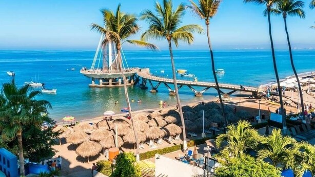 Un resort sulla spiaggia con una passeggiata alberata da palme, un pontile di legno e un'area salotto con tetto di paglia.