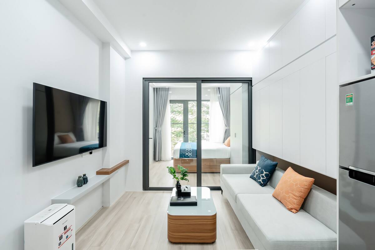 A modern, well-lit room with a flat-screen TV, a coffee table, a sofa with cushions, a refrigerator, and a sliding glass door leading to a balcony with a bed.