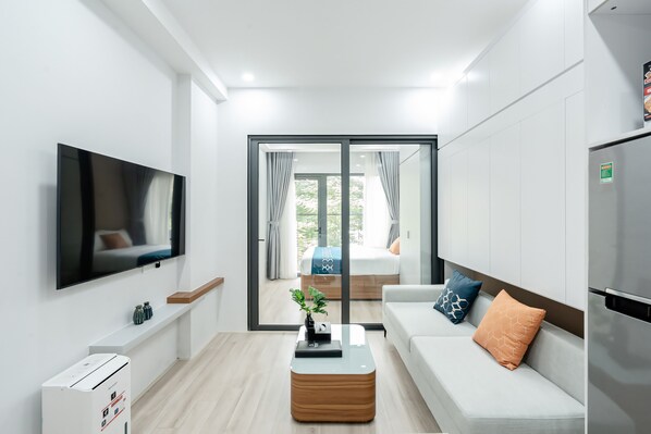 A modern, well-lit room with a flat-screen TV, a coffee table, a sofa with cushions, a refrigerator, and a sliding glass door leading to a balcony with a bed. - FLESTA Nui Truc Serviced Apartment & Hotel (Hanoi)