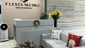 A modern serviced apartment with a reception desk, a grey sofa, a red plaid ottoman, and a large window with a view of the city. - FLESTA Nui Truc Serviced Apartment & Hotel (Hanoi)