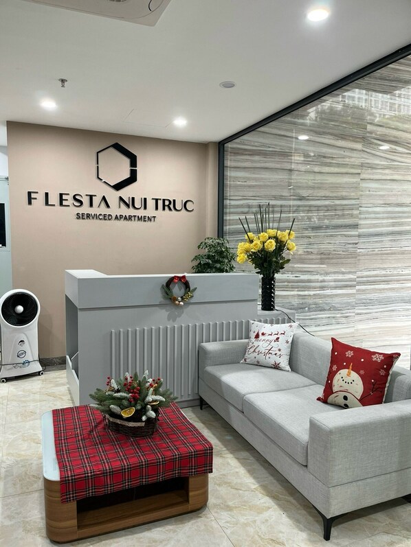 A modern serviced apartment with a reception desk, a grey sofa, a red plaid ottoman, and a large window with a view of the city. - FLESTA Nui Truc Serviced Apartment & Hotel (Hanoi)