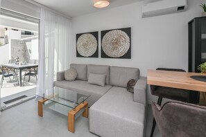 Un salon moderne avec un canapé gris, une table basse en verre et une table à manger en bois. Deux unités de climatisation sont fixées au mur, et un pot de fleurs se trouve sur la droite.