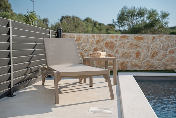 Ein Liegebereich am Pool mit einer einzelnen, beige gestreiften Liege, einem kleinen Tisch mit einem Handtuch und einer Steinwand im Hintergrund.