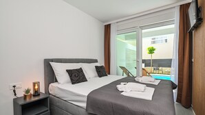 A modern hotel room with a large bed, a bedside table with a lamp, and a view of a swimming pool. - Smash A - Apartments with private pool  (Novalja)