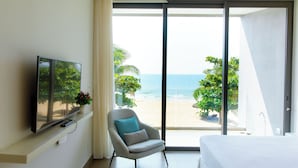 Habitación de hotel con cama, televisor sobre un mueble, sillón con cojín y un amplio ventanal corredizo con vista a la playa.