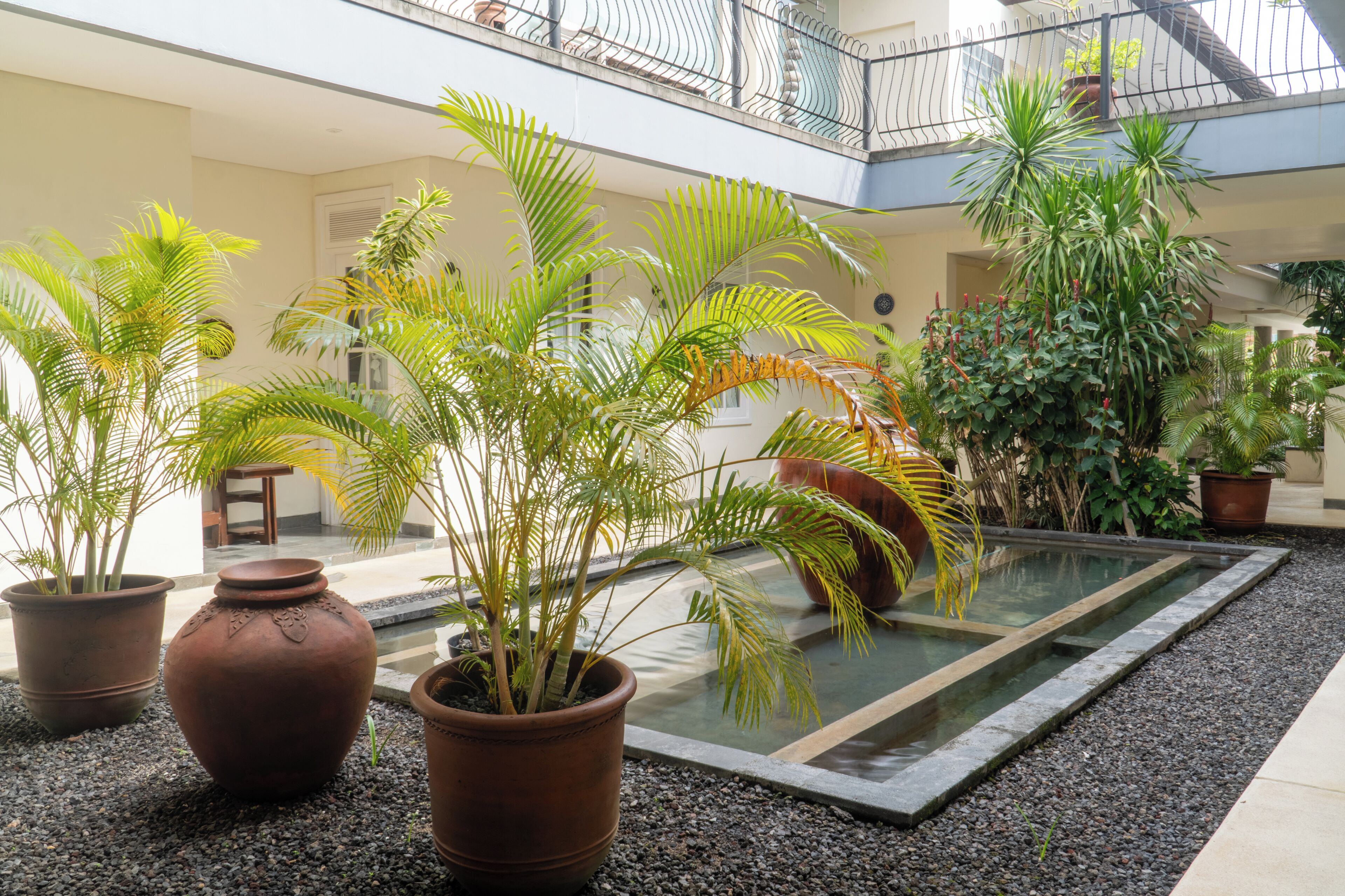 A spacious indoor atrium with a central water feature, surrounded by potted plants and a balcony with a railing.