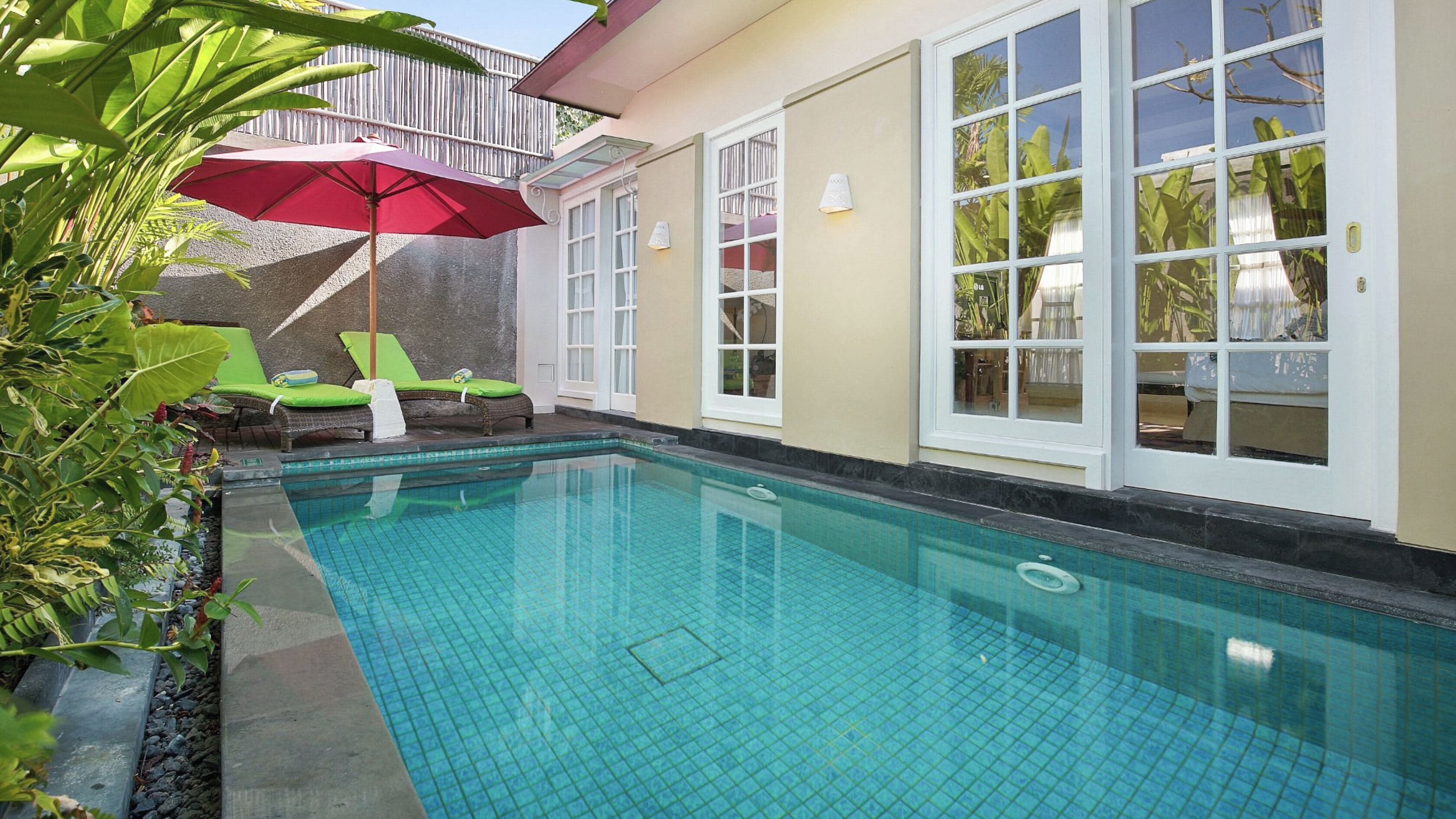 Ein Swimmingpool mit Fliesenboden, einem roten Sonnenschirm und zwei grünen Liegestühlen.