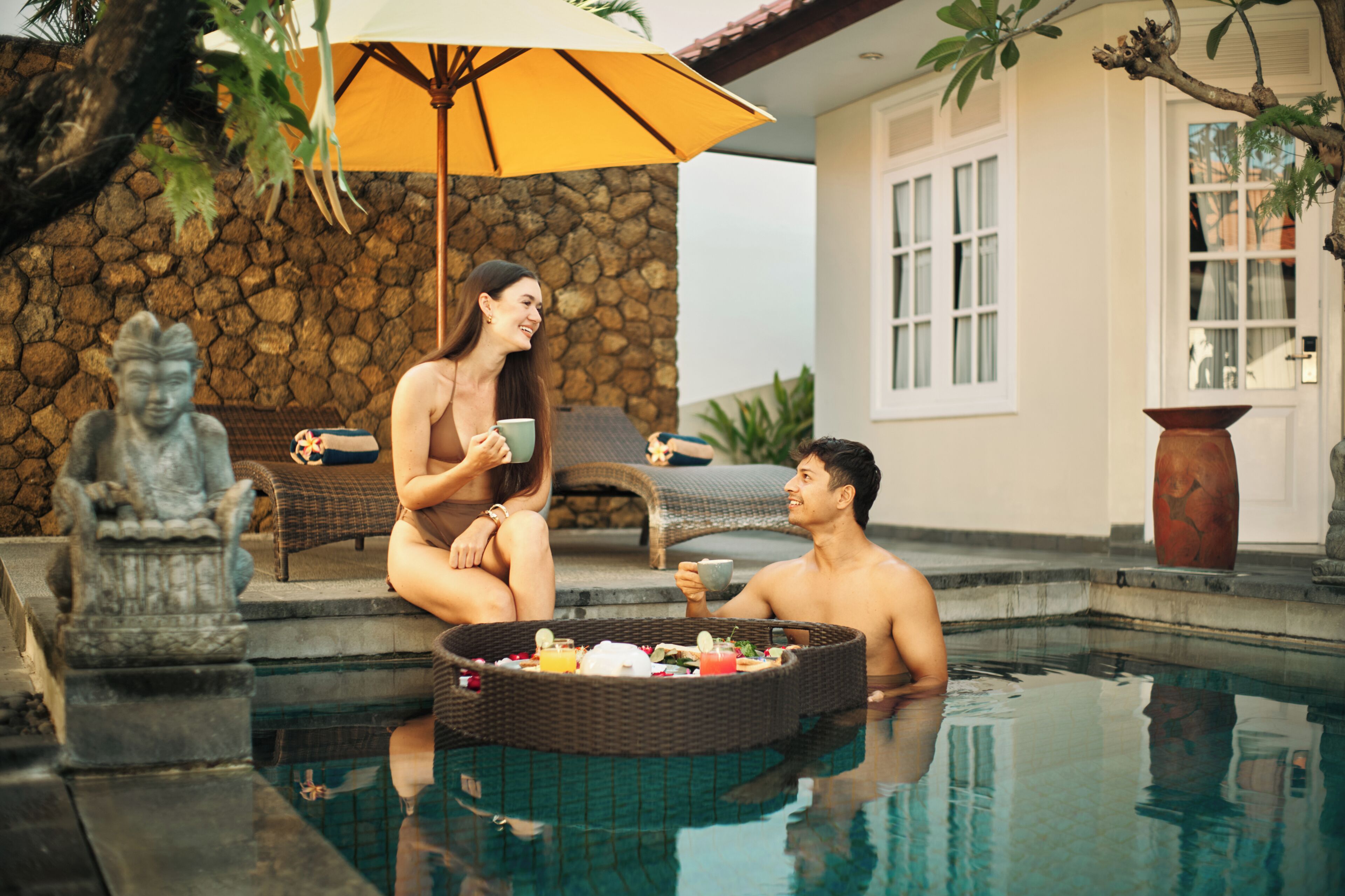 A poolside setting with a stone wall, a statue, and a wicker table with food and drinks.