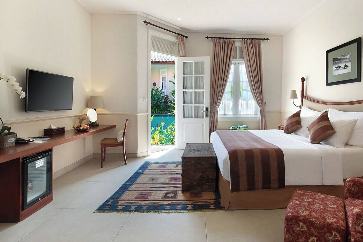 A hotel room with a bed, a desk with a television, a chair, a small table, a nightstand, and a window with curtains.