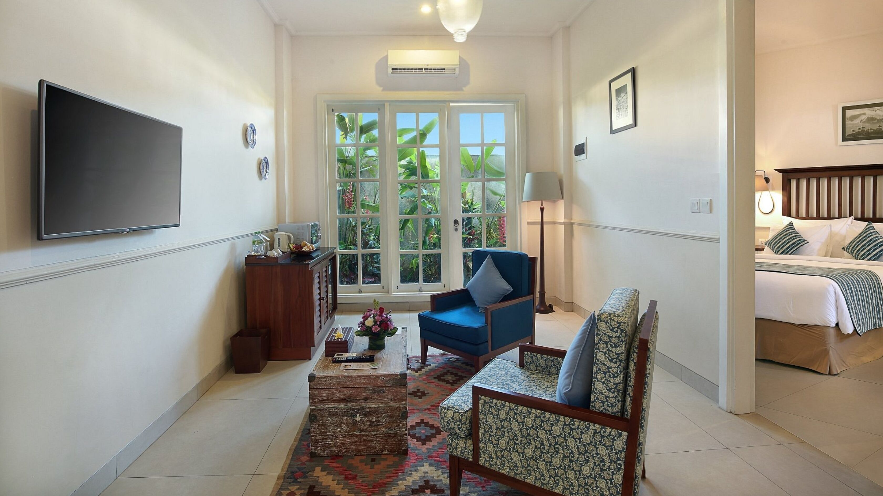 A hotel room with a TV, a desk with a lamp, a chair, a bed, and a view of greenery through the windows.