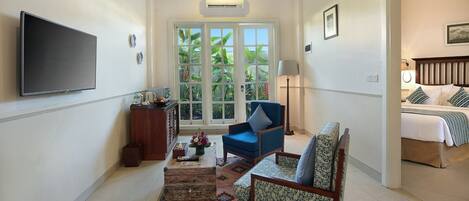 A hotel room with a TV, a desk with a lamp, a chair, a bed, and a view of greenery through the windows.