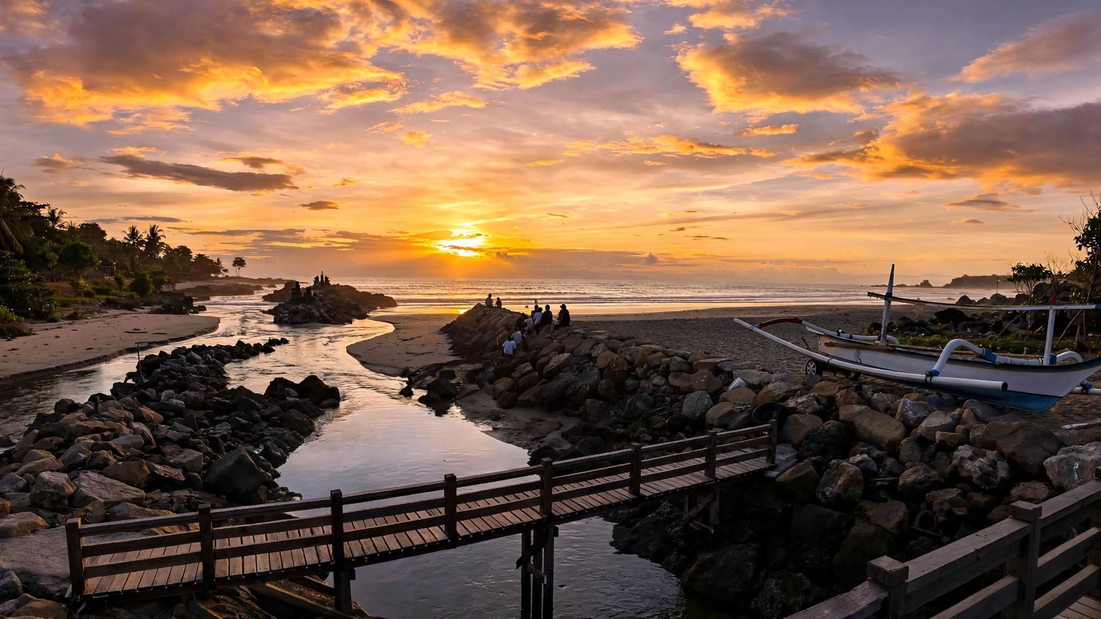A wooden bridge over a rocky riverbed leading to a beach with a sunset in the background.