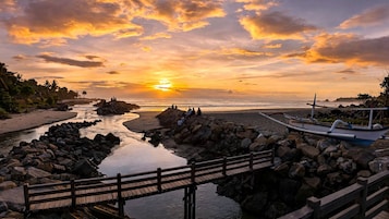A wooden bridge over a rocky riverbed leading to a beach with a sunset in the background.