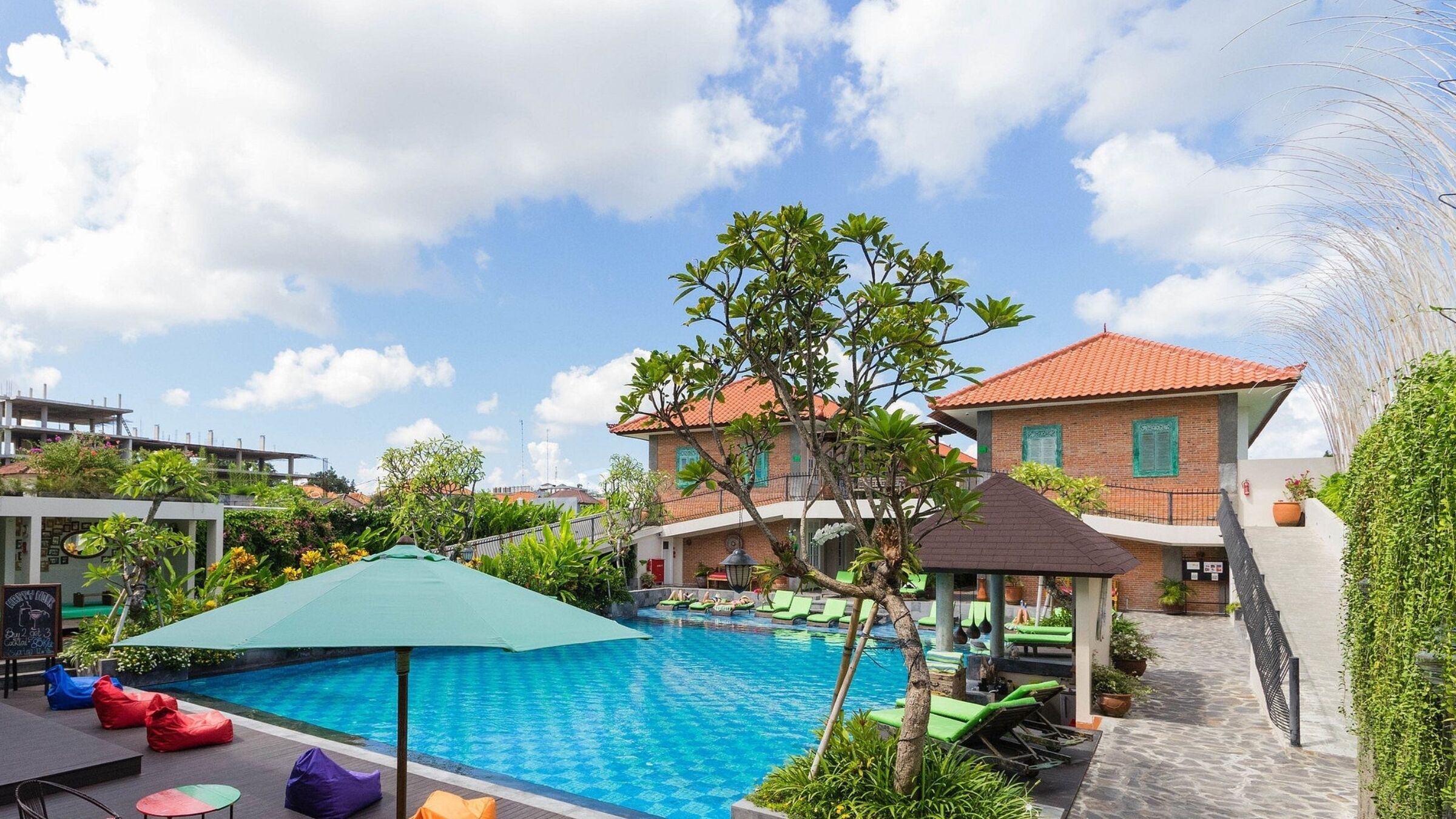 A swimming pool with lounge chairs and a covered seating area, surrounded by greenery and a building with a tiled roof.