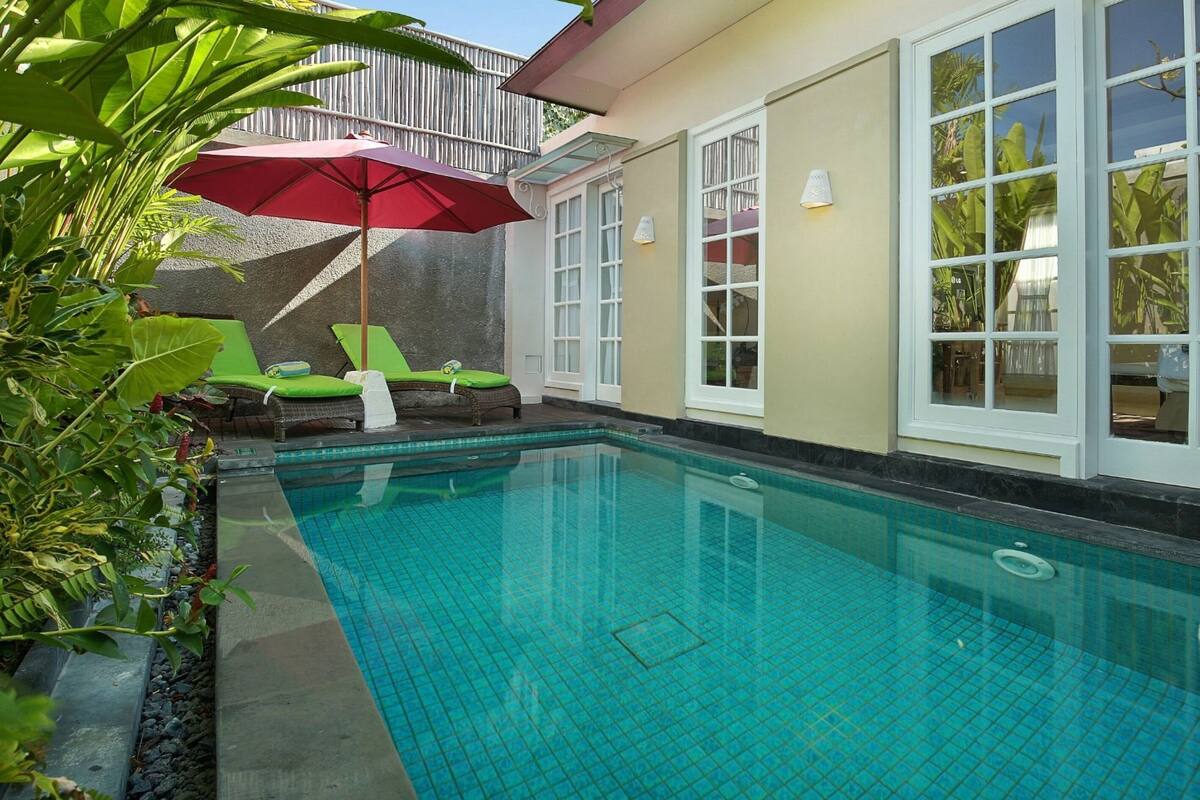 A swimming pool with tiled flooring, a red umbrella, and green lounge chairs.