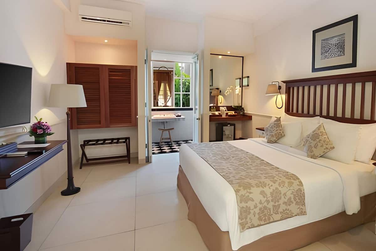 A hotel room with a bed, bedside tables, a desk with a lamp, a TV, and a bathroom visible through an open door.