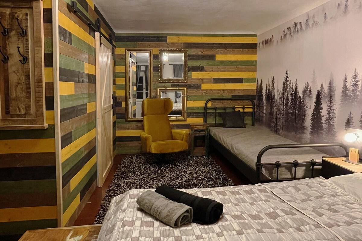Une pièce comprenant un lit, une chaise, un miroir et un mur orné d’une fresque représentant une forêt.