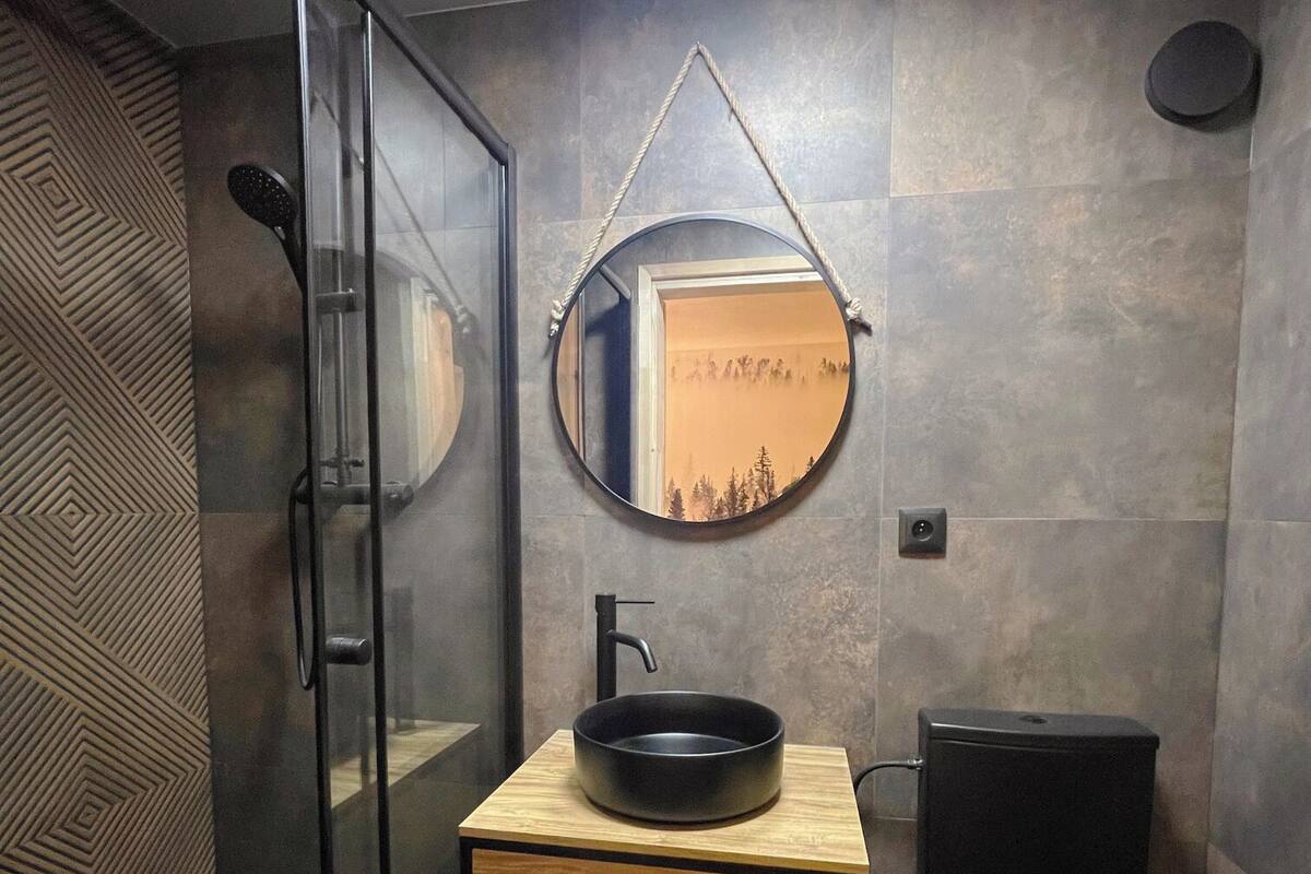 Une salle de bain moderne avec une douche à parois de verre, un meuble-lavabo en bois avec un vasque noire et un miroir rond accroché au mur.