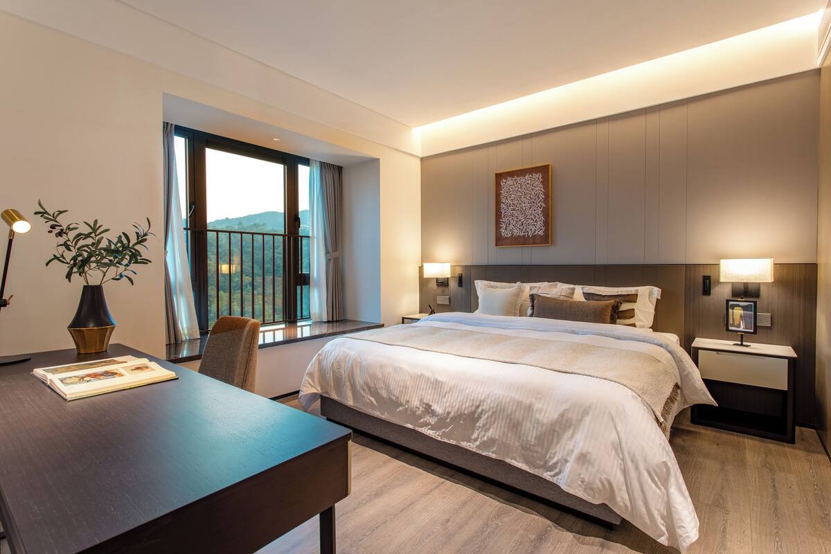 Ein modernes Hotelzimmer mit einem großen Bett, Nachttischen mit Lampen, einem Schreibtisch mit Stuhl, einer Topfpflanze und einem Fenster mit Blick ins Freie.