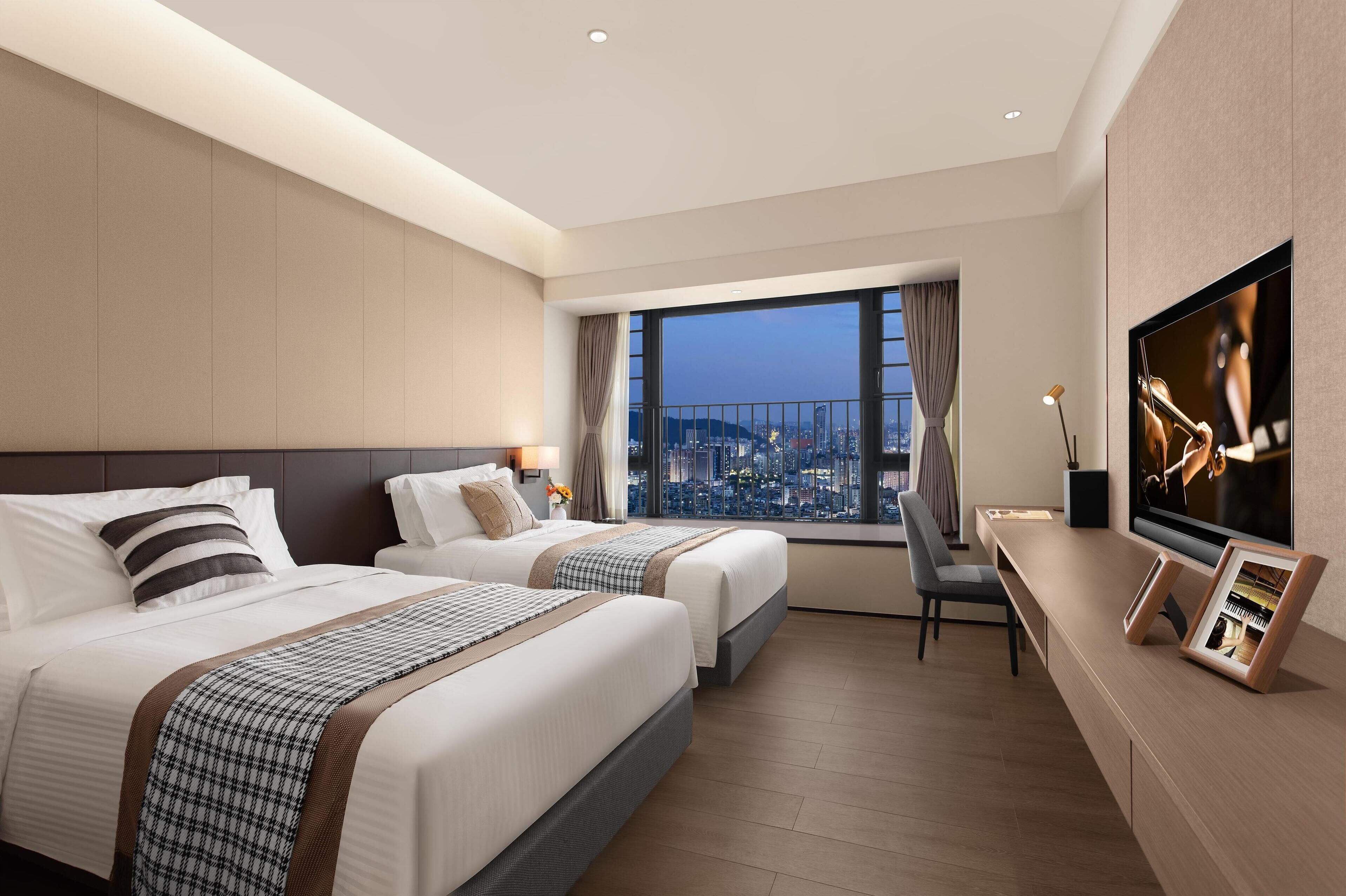 A hotel room with two beds, a TV, a desk, and a large window with a city view.