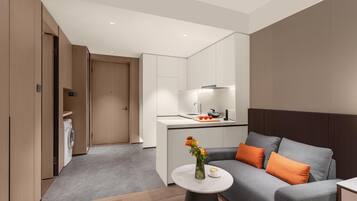Una habitación de hotel moderna con un sofá gris, cojines naranjas, una pequeña mesa redonda con un florero y una zona de cocina con fregadero y estufa.
