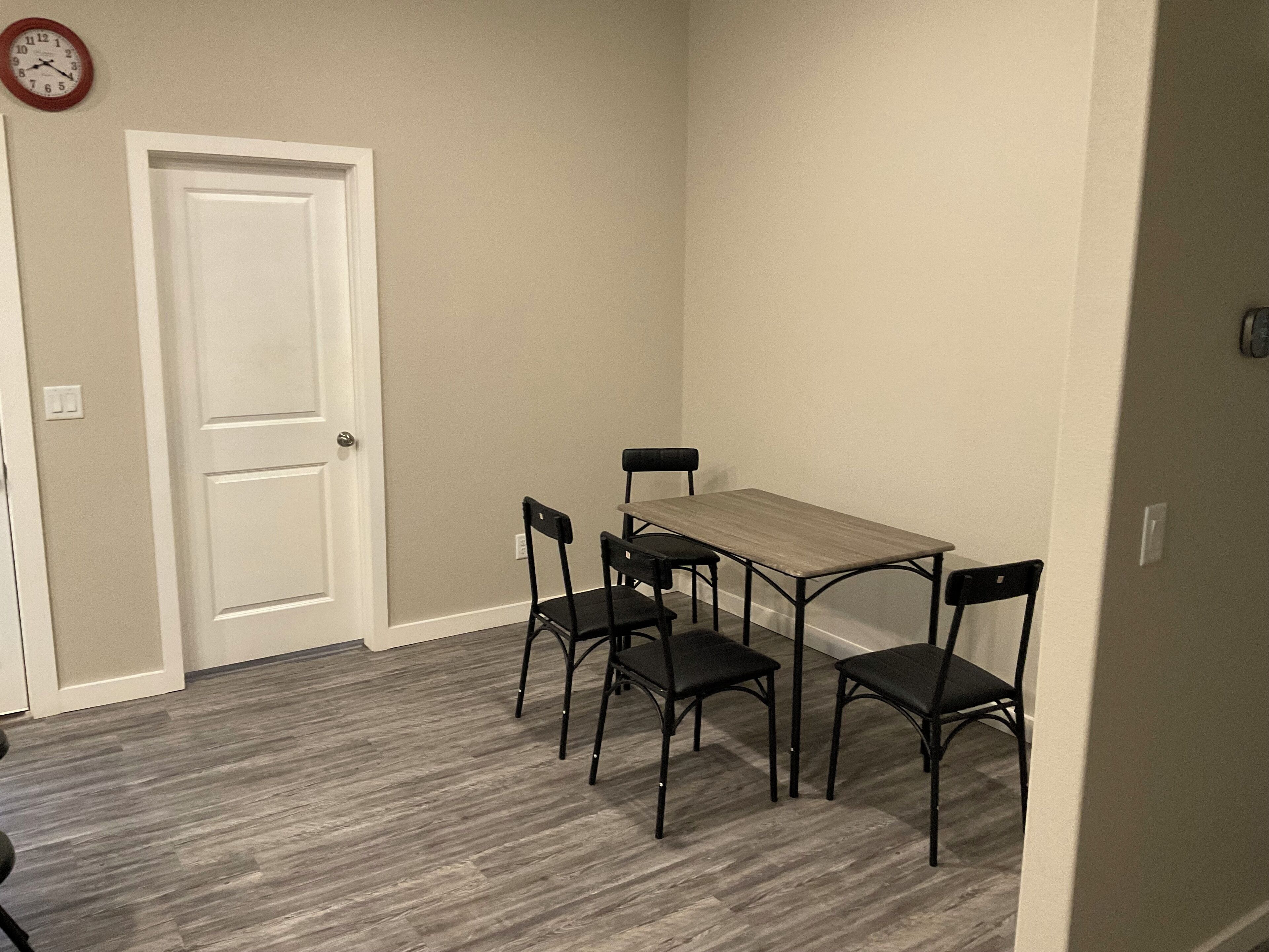 Una habitación pequeña con una mesa de madera y cuatro sillas, una puerta blanca y un reloj en la pared.