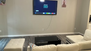 Una sala de estar con un televisor de pantalla plana montado en la pared, un sofá blanco, un pequeño reposapiés y una mesa de centro negra.